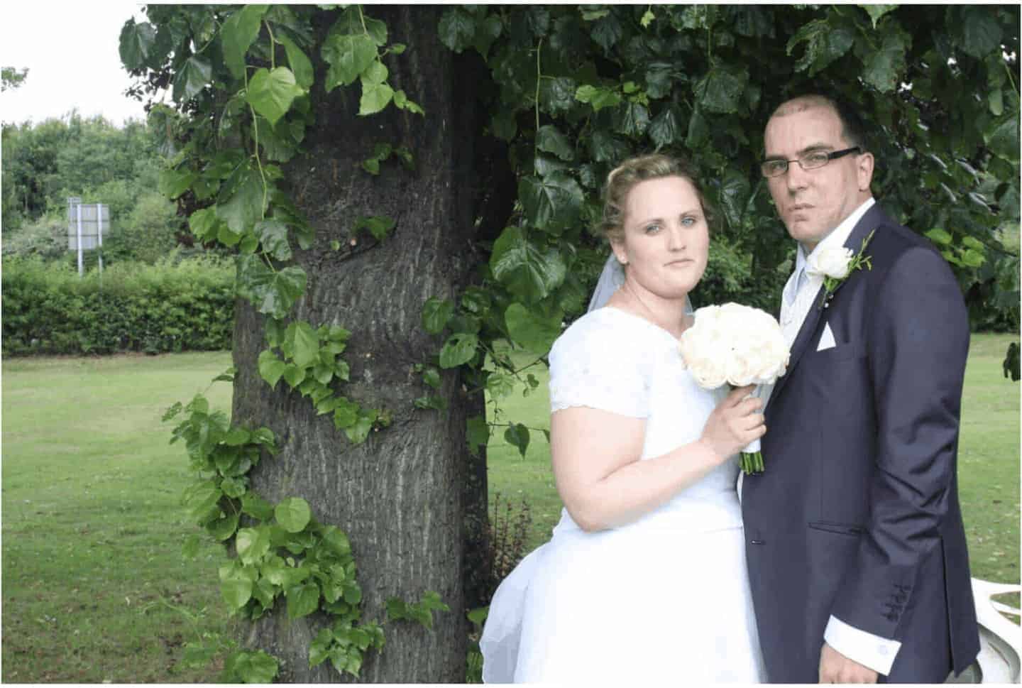 angela and john milnes wedding photo. a bride and groom standing bu a tree and holding flowers.