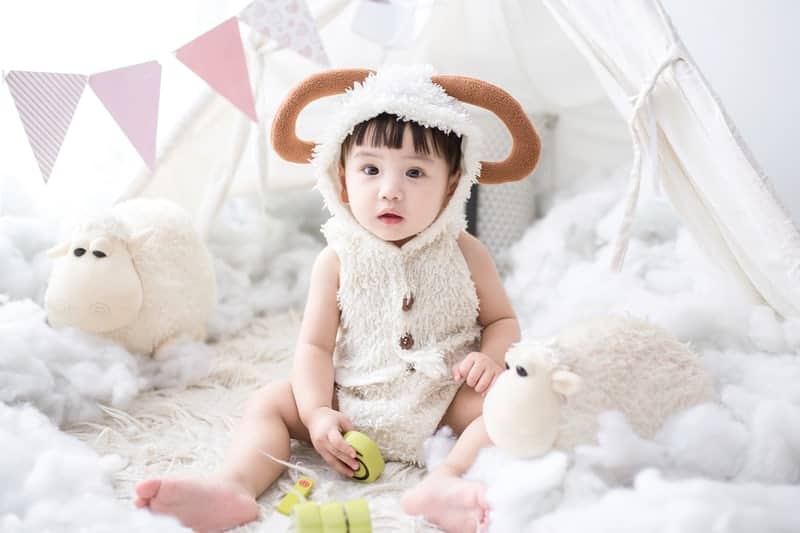 Toddler girl wearing cute sheep costume with stuffed animals nearby.
