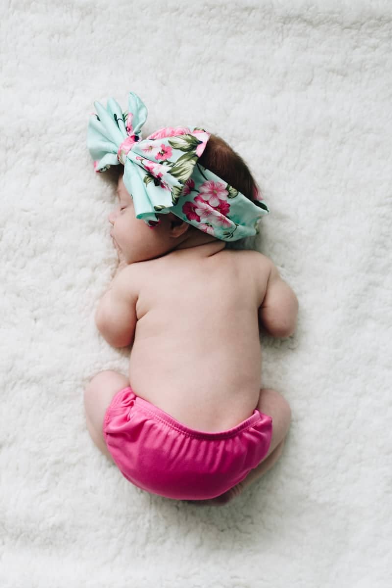 Newborn baby curled up on stomach wearing flowered headband and pink diaper cover.