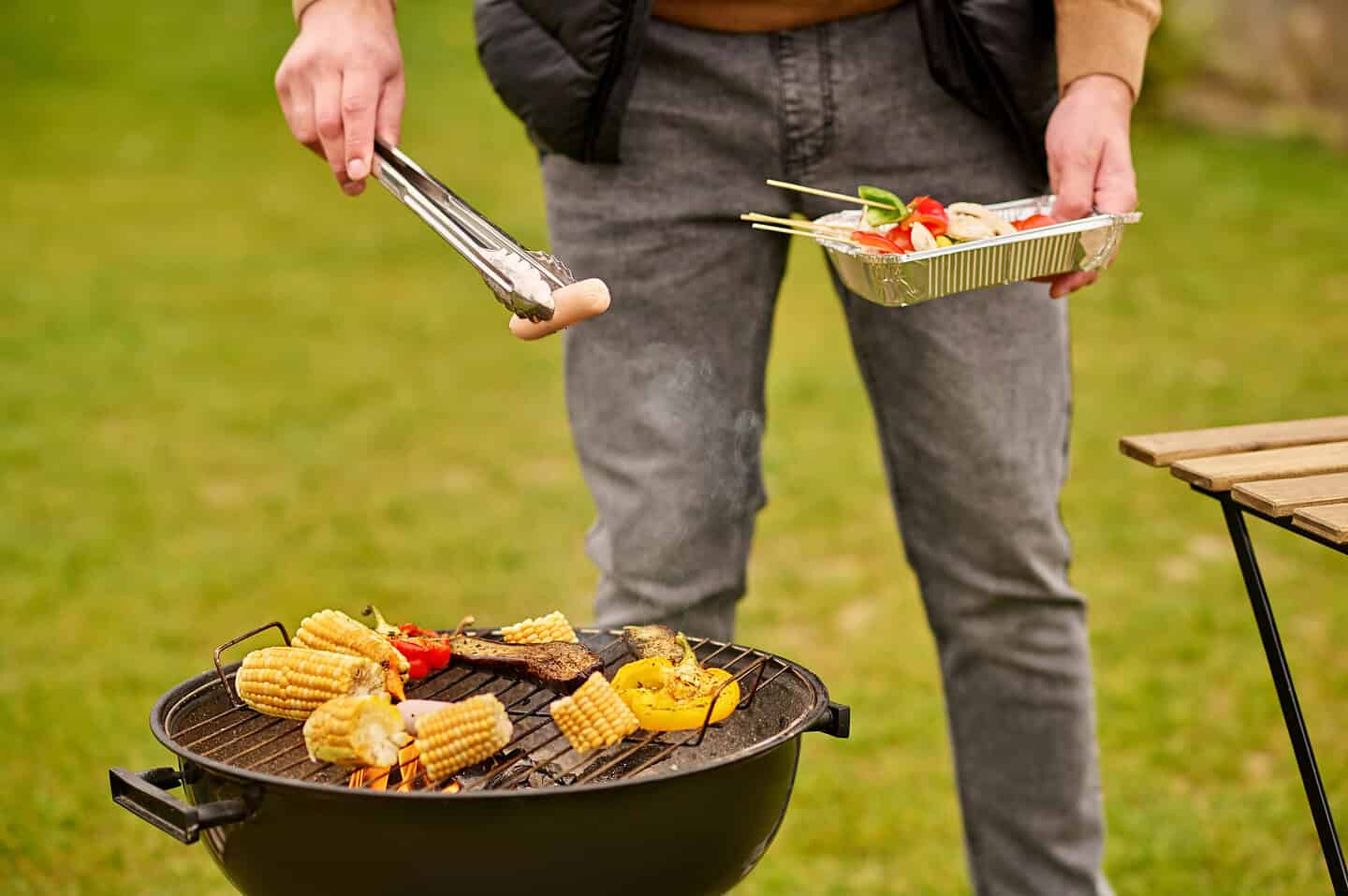 Picnic, barbecue. Male hand holding tongs with sausage over vegetables on barbecue on green lawn in garden in autumn day, no face